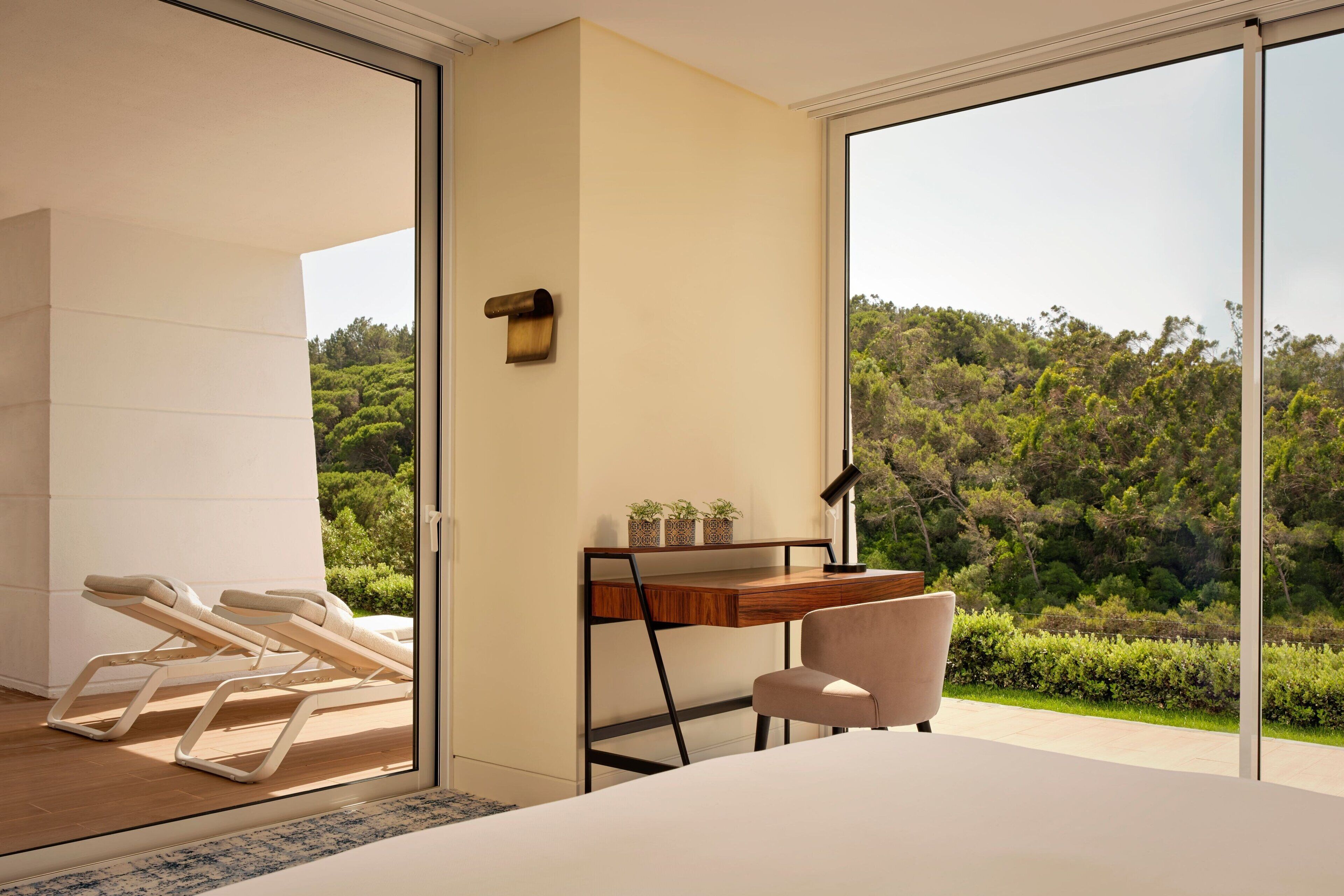A modern bedroom with a large window, a wooden desk, a chair, and two lounge chairs.