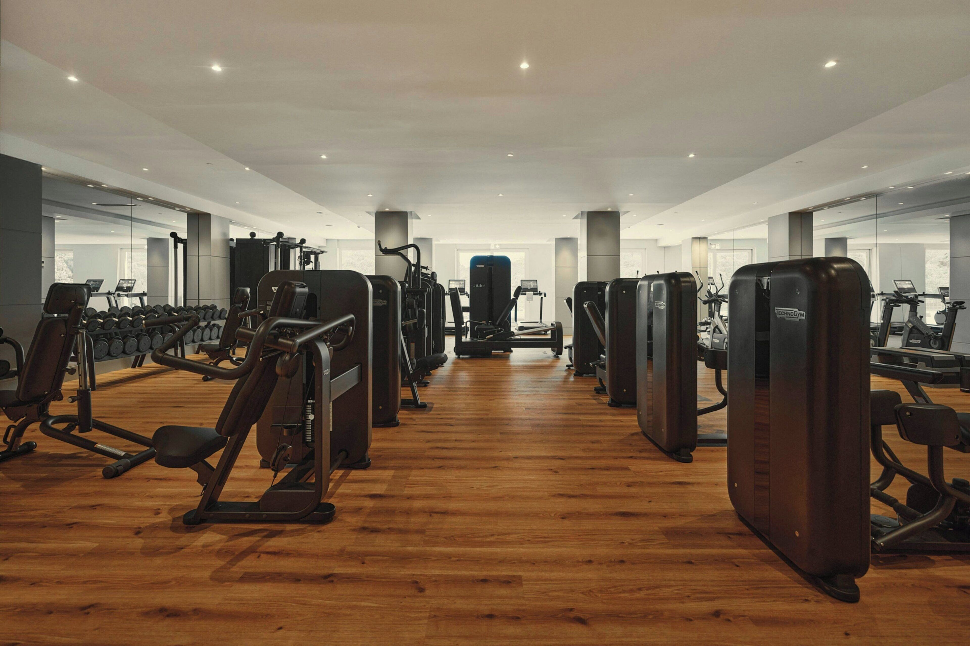 Un gimnasio con diversas máquinas de ejercicio, suelo de madera y paredes con espejos.