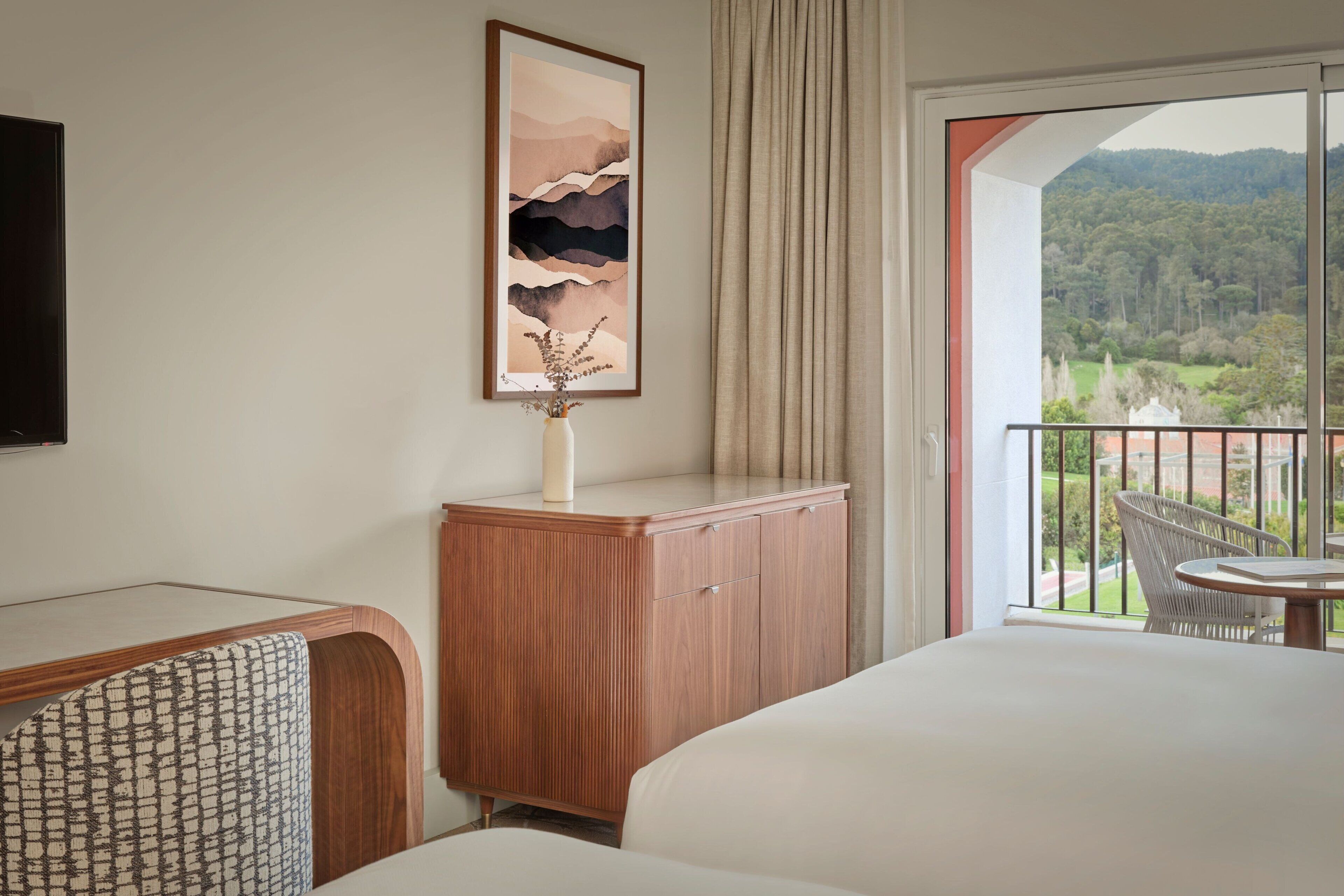 Habitación de hotel con cama, cómoda de madera, obra de arte enmarco, jarrón con plantas secas, silla y balcón con vista a un paisaje boscoso.
