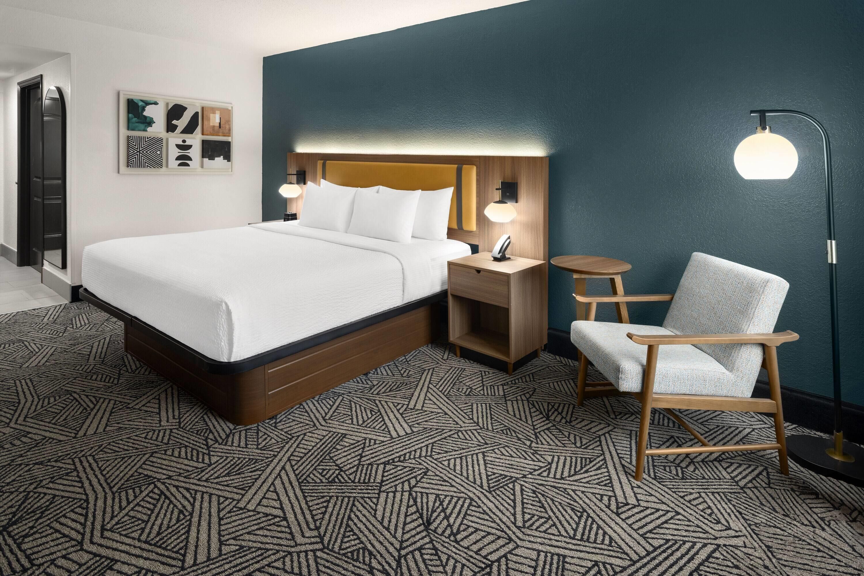 Una habitación de hotel con una cama grande, una mesita de noche, una silla y una lámpara de pie.