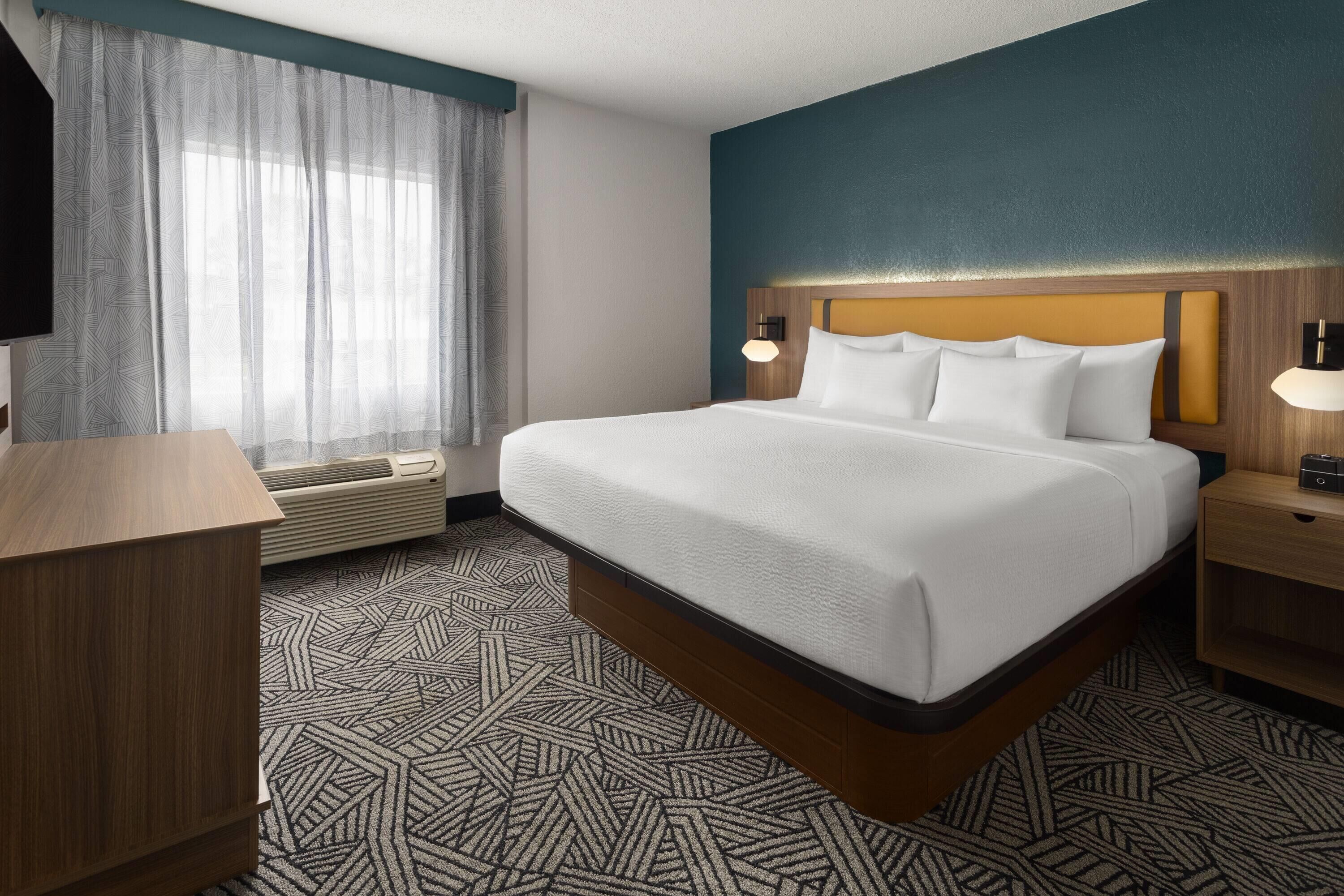 Une chambre d’hôtel avec un grand lit, une table de chevet en bois, un bureau, une fenêtre avec des rideaux légers et un téléviseur fixé au mur.