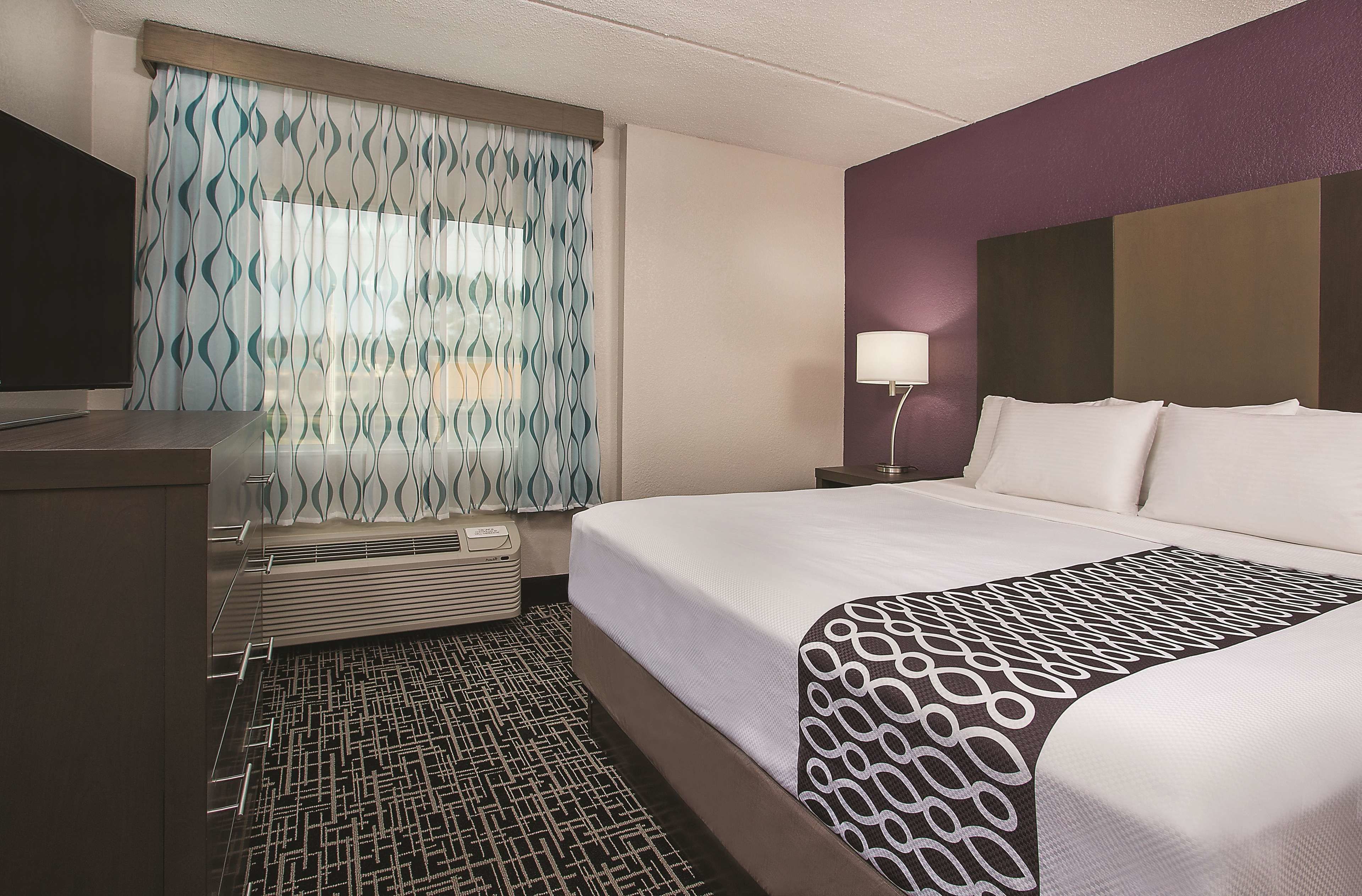 A hotel room with a bed, a desk, a lamp, and a window with patterned curtains.