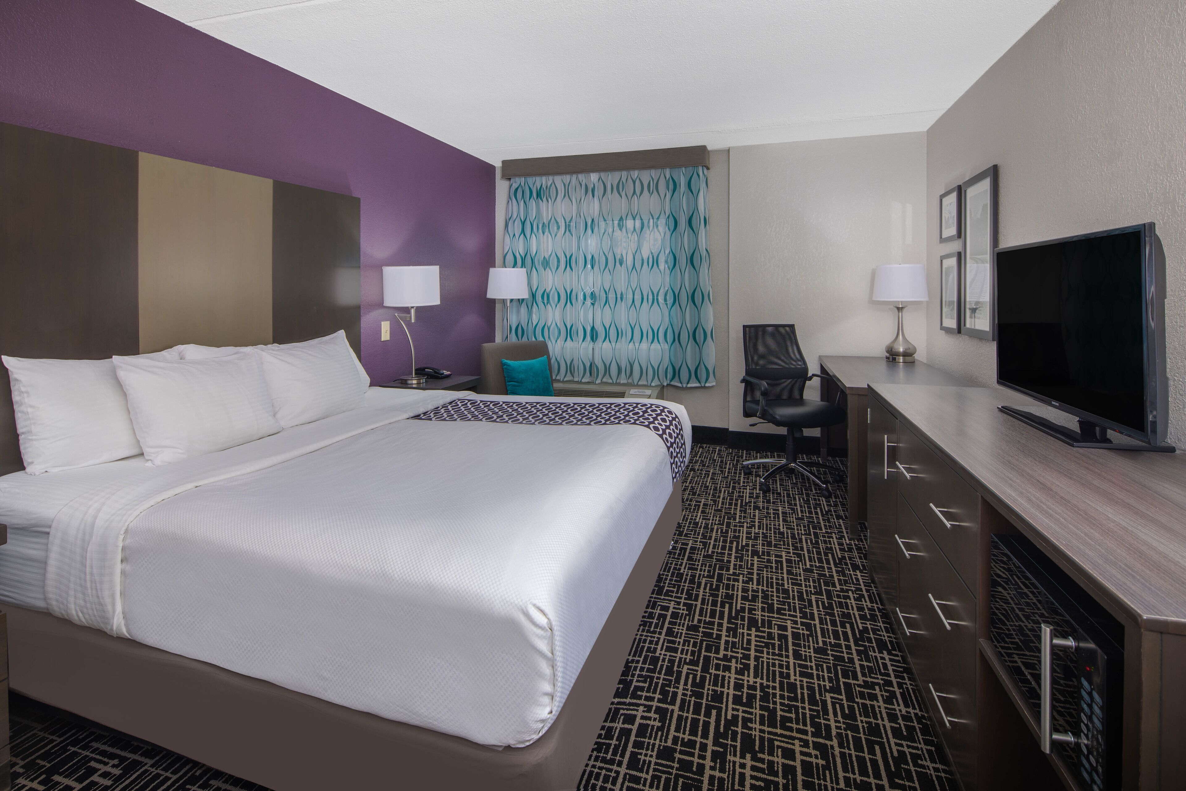 A hotel room with a large bed, a desk with a television, two chairs, and a patterned carpet.