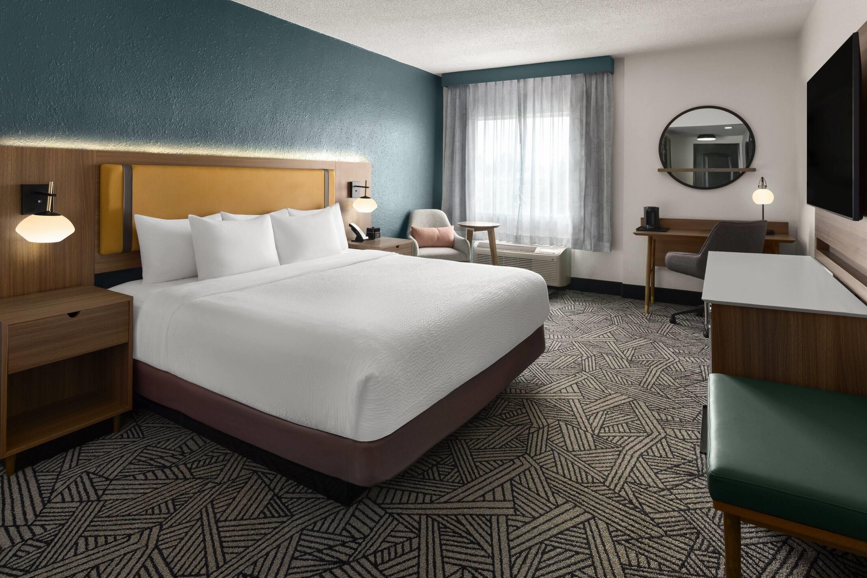 Une chambre d’hôtel avec un grand lit, un bureau, une chaise, une petite table, un téléviseur et une fenêtre avec des rideaux.