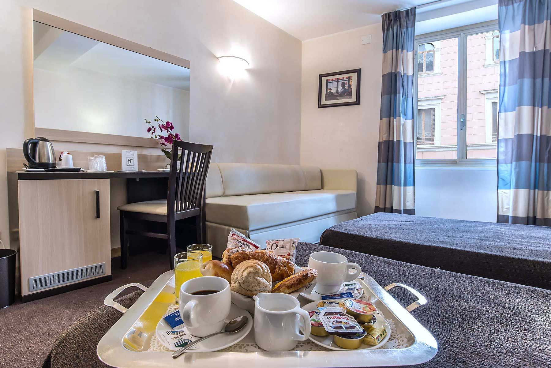 Habitación de hotel con cama, sofá, escritorio con silla y bandeja de desayuno con café, jugo, croissants y pastelería.