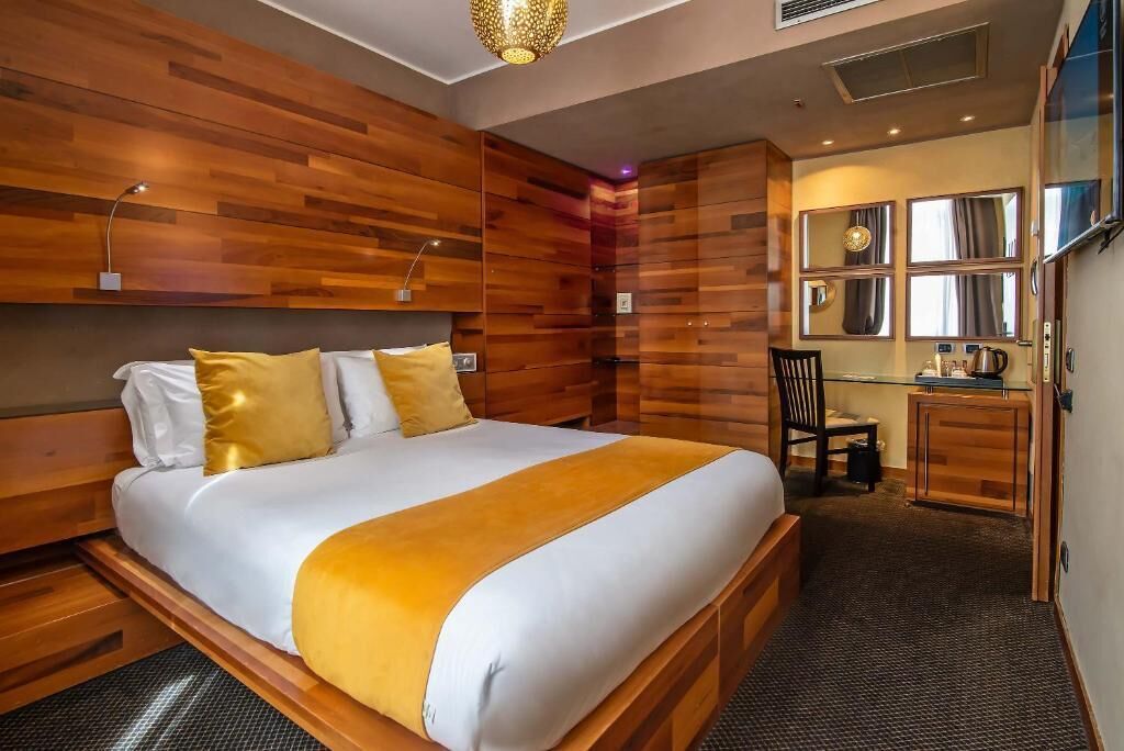 Een moderne hotelkamer met een houten hoofdeinde, een groot bed met wit en geel beddengoed, een bureau met stoel en een spiegel boven het bureau.