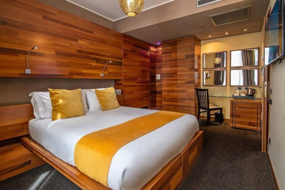 Une chambre d’hôtel moderne avec une tête de lit en bois, un grand lit avec une literie blanche et jaune, un bureau avec une chaise et un miroir au-dessus du bureau.