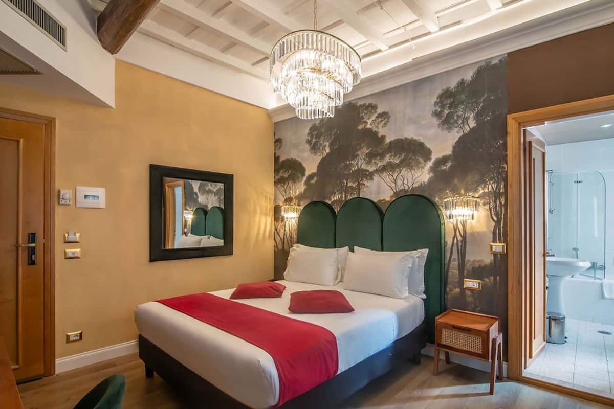 Une chambre à coucher avec un lit, un lustre, un tableau et une salle de bain visible à travers une porte ouverte.