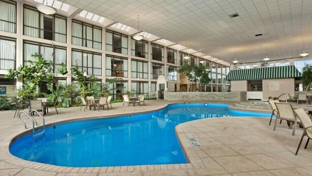 An indoor swimming pool with multiple lanes, surrounded by lounge chairs and tables, and a large building with numerous windows and a green awning. - Days Inn by Wyndham Columbus Airport (Columbus)