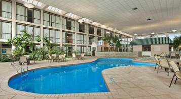 An indoor swimming pool with multiple lanes, surrounded by lounge chairs and tables, and a large building with numerous windows and a green awning.