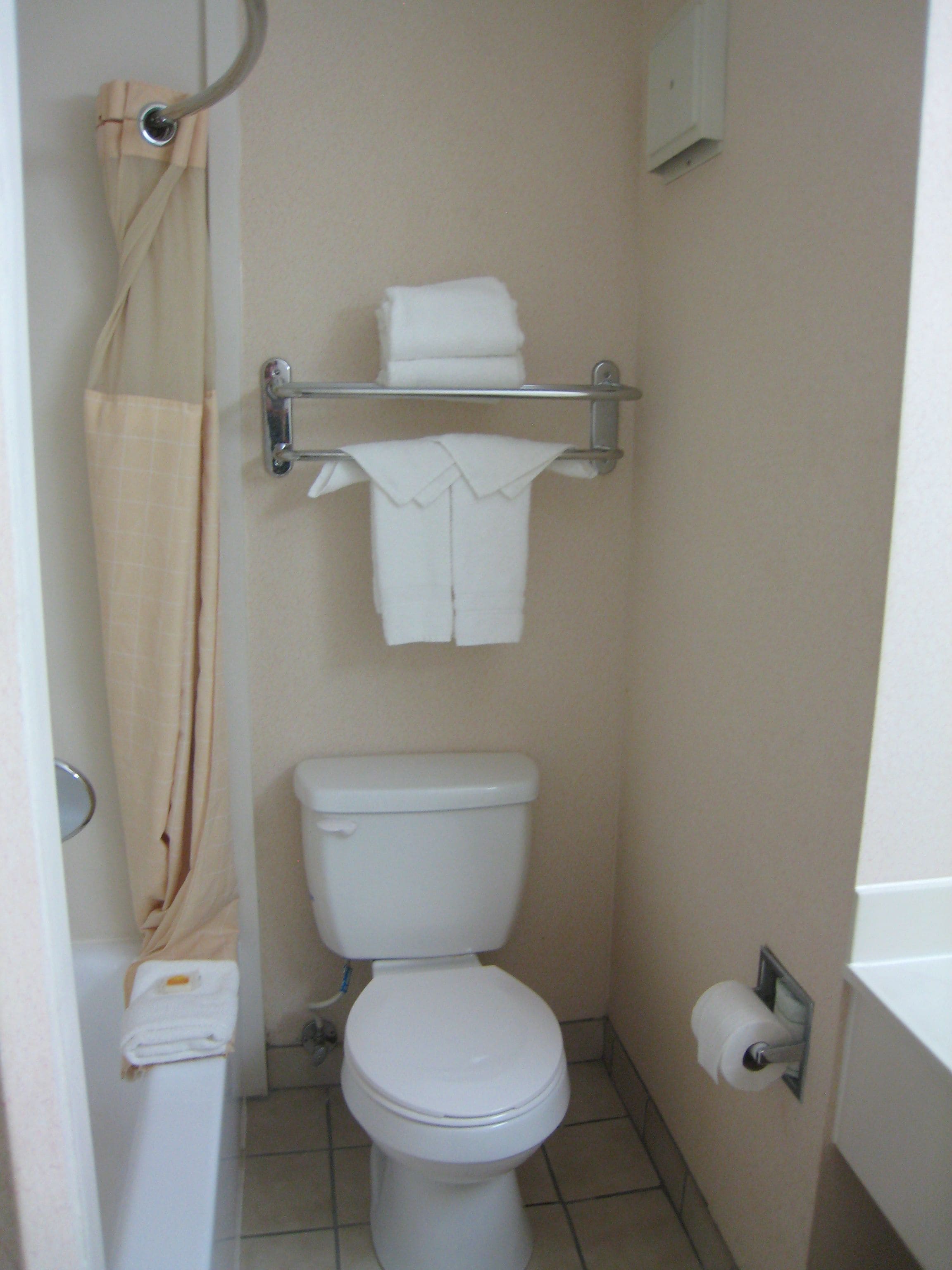 Un baño pequeño con inodoro, cortina de ducha, toallero y una toalla doblada, además de un dispensador de papel higiénico.
