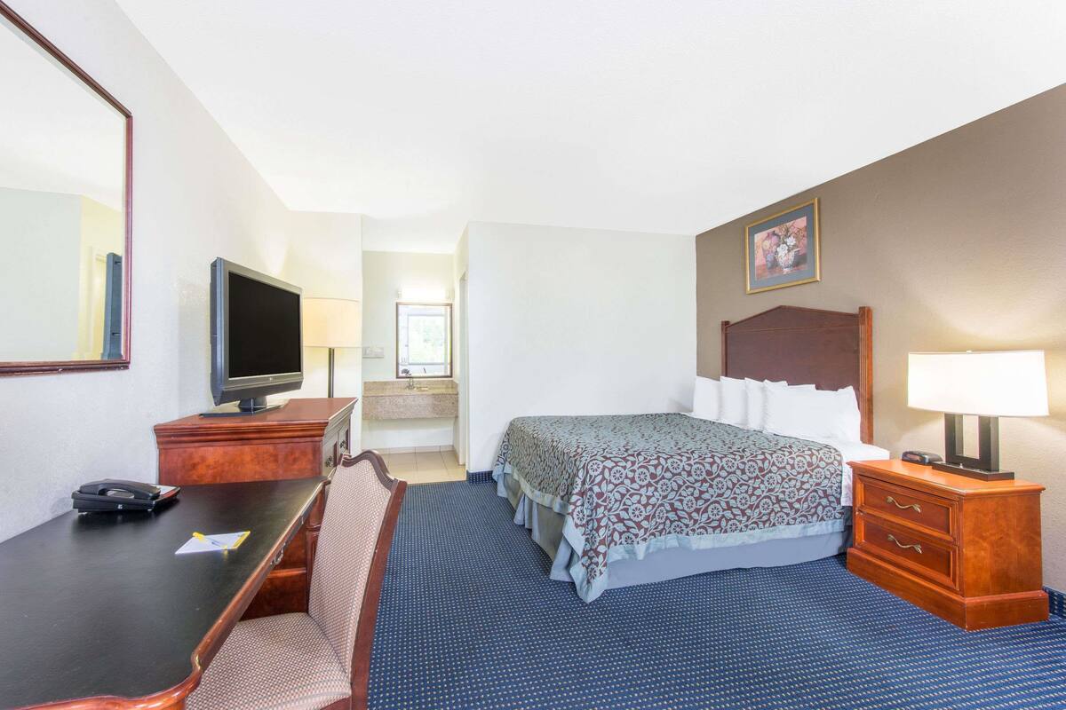 A hotel room with a bed, a desk with a telephone, a television, a mirror, a bedside table with a lamp, and a bathroom visible in the background.