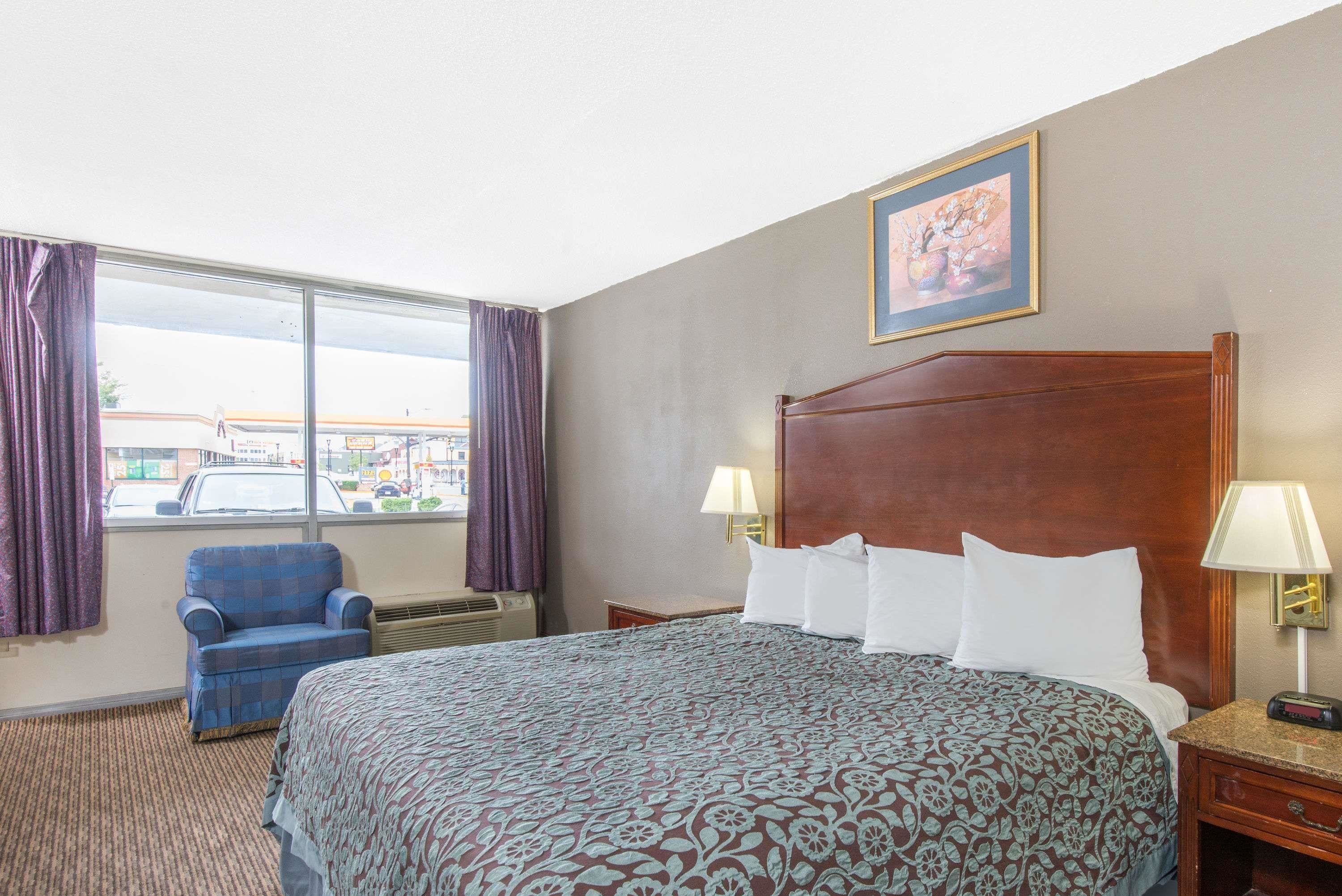 A hotel room with a large bed, a chair, a window with curtains, and a painting on the wall.