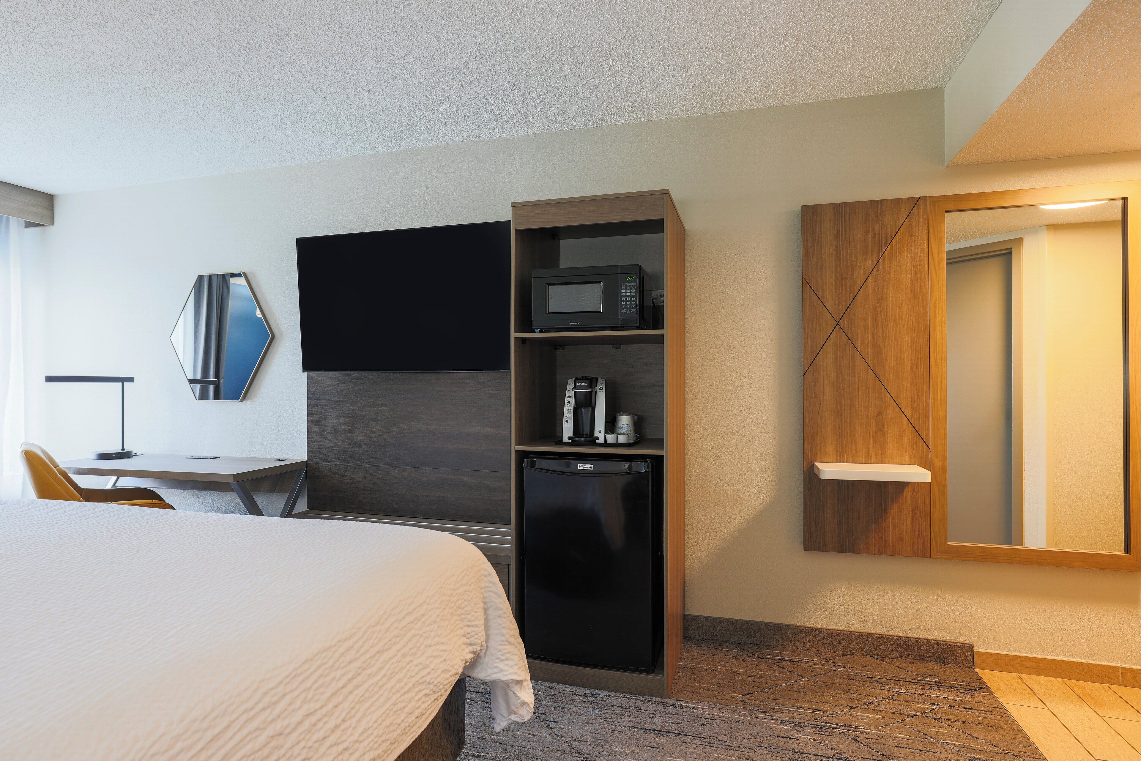 A hotel room with a bed, a desk, a flat-screen TV, a microwave, a coffee maker, and a minibar.