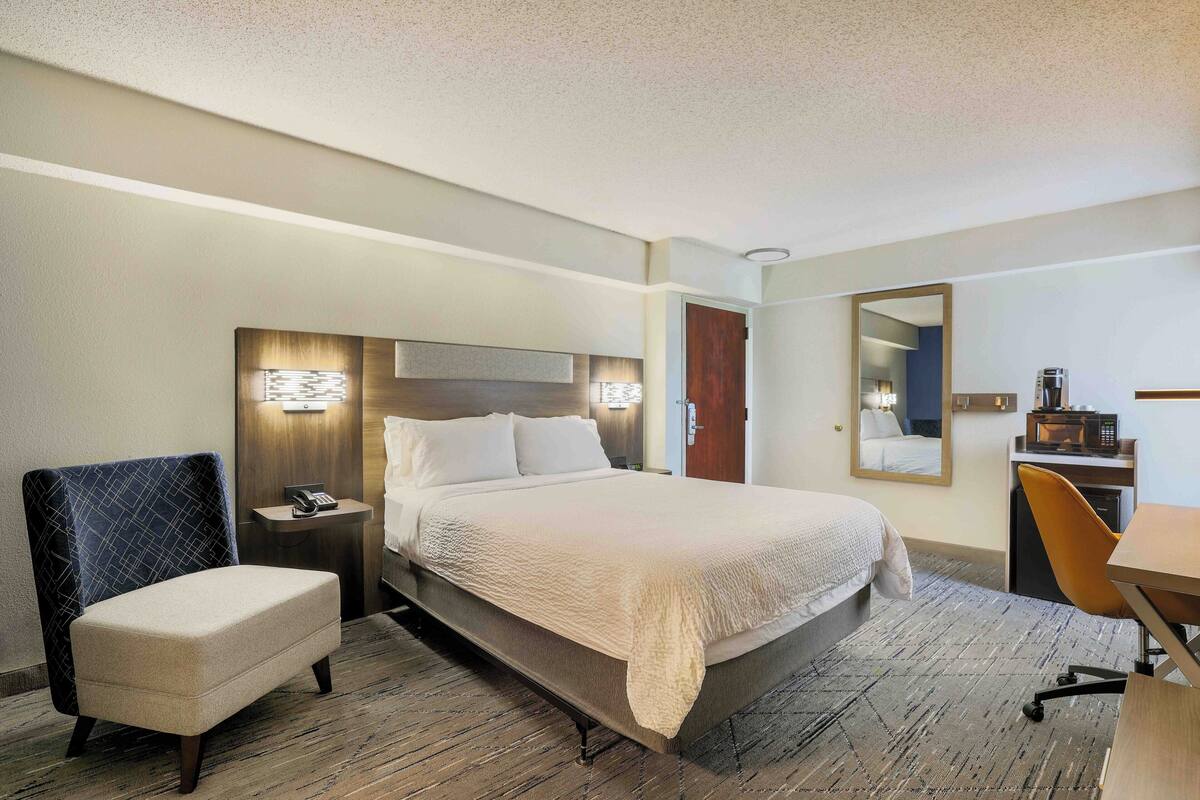 A hotel room with a large bed, a chair, a desk, a microwave, and a mirror.