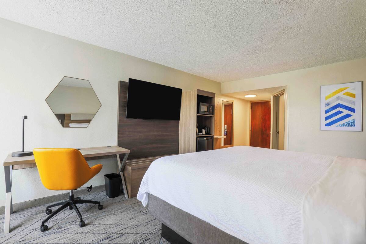 A hotel room with a bed, a desk with a chair, a flat-screen TV, a microwave, and a door leading to another room.