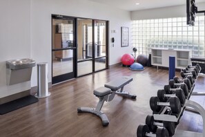 Academia com equipamentos de ginástica, um bebedouro e vista para o exterior através de uma porta de vidro.