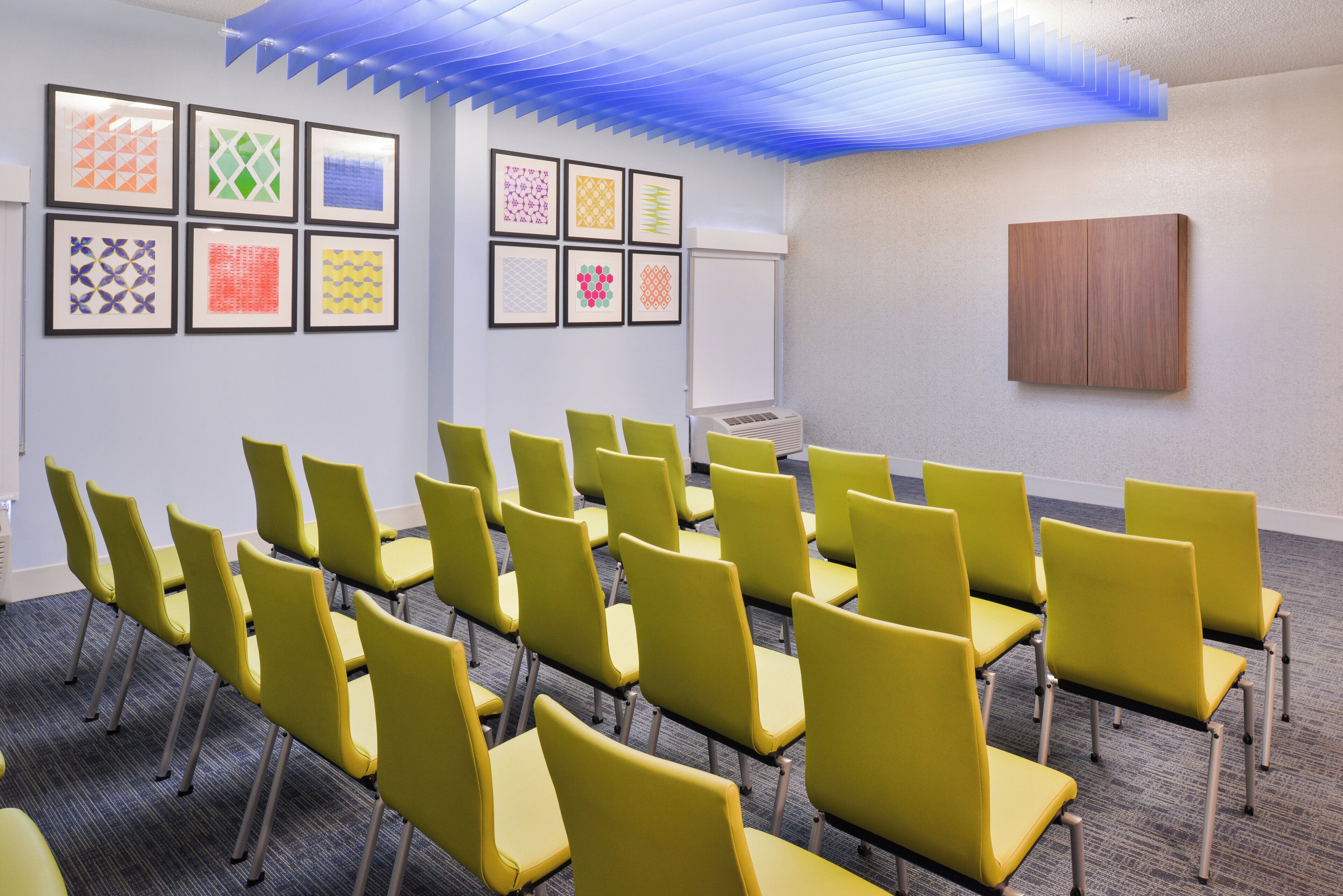 Un salón de conferencias con hileras de sillas amarillas, una pizarra y obras de arte geométricas enmarcadas en las paredes.