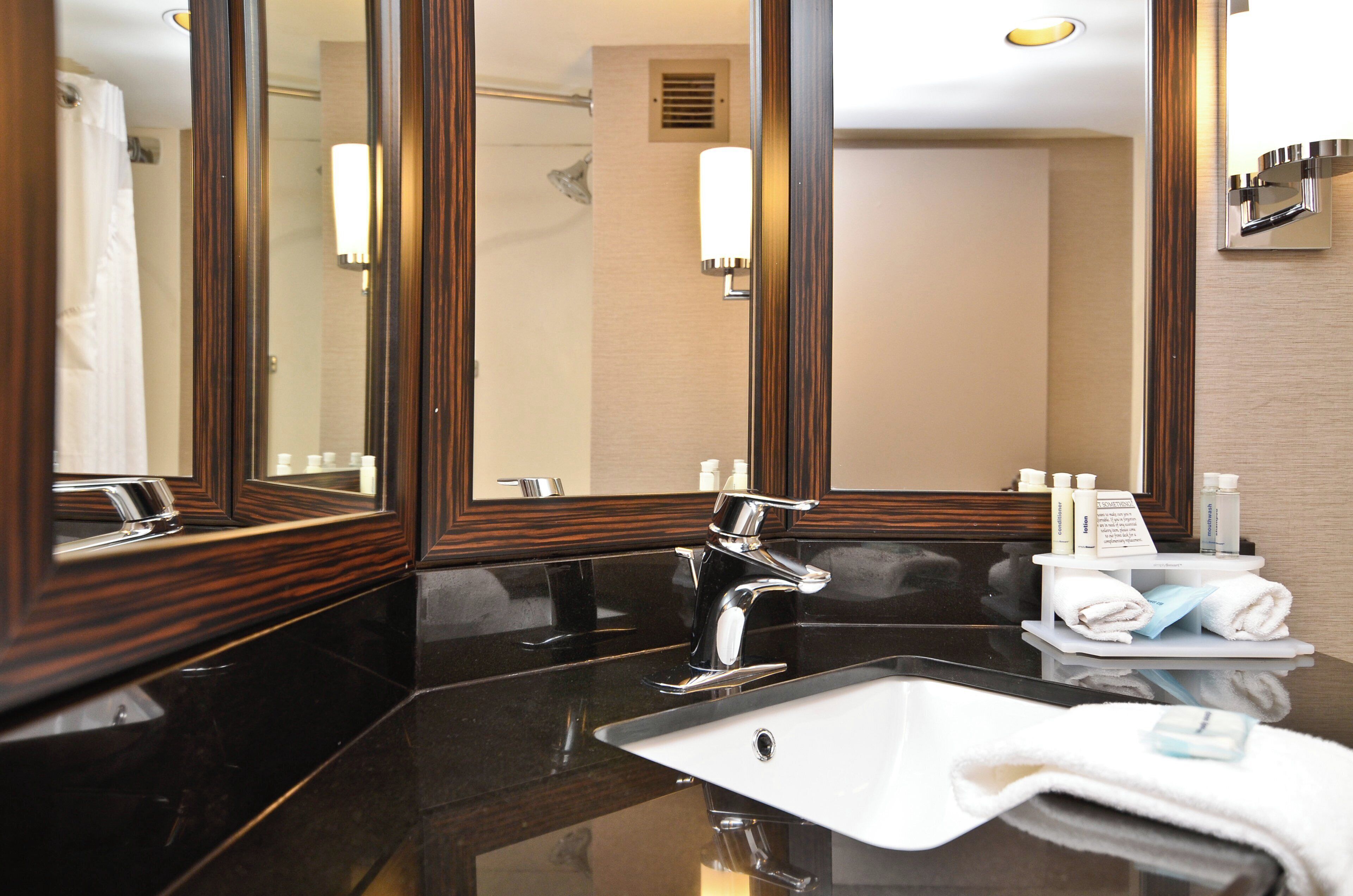 A hotel bathroom with a large mirror, a modern sink, and toiletries on a shelf.