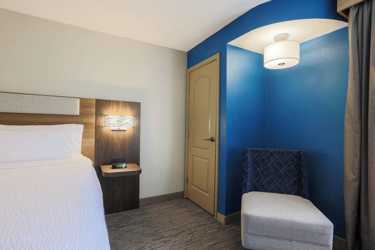 A hotel room with a bed, bedside table, lamp, chair, and a door.
