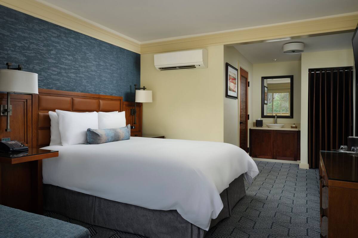 A hotel room with a large bed, a bedside table with a lamp, a desk with a telephone, a bathroom with a sink and mirror, and a blue accent wall.