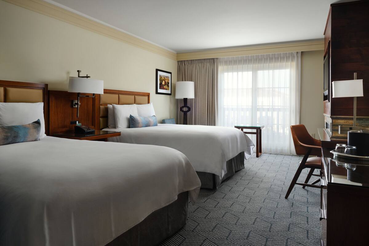 A hotel room with two beds, a desk, a chair, a lamp, and a painting on the wall.