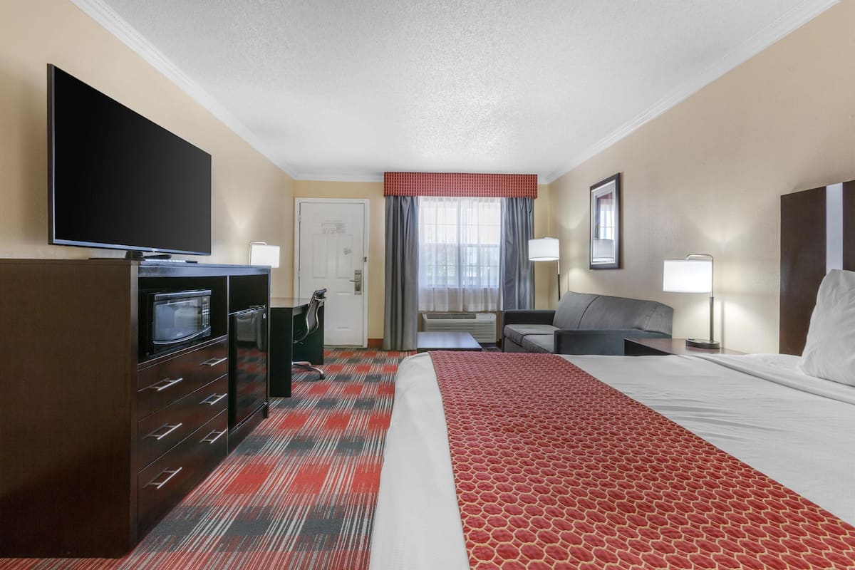 Habitación de hotel con cama, televisor, escritorio con microondas, silla, sofá y ventana con cortinas.