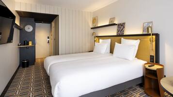 A modern hotel room with a large bed, a bedside table, a wall-mounted shelf with artwork, and a patterned floor.