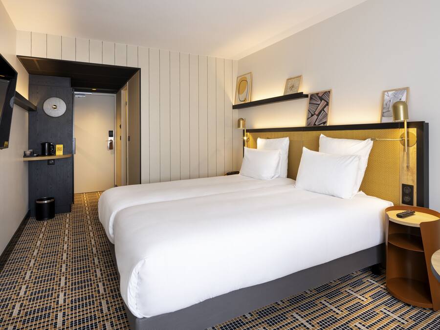 A modern hotel room with a large bed, a bedside table, a wall-mounted shelf with artwork, and a patterned floor.