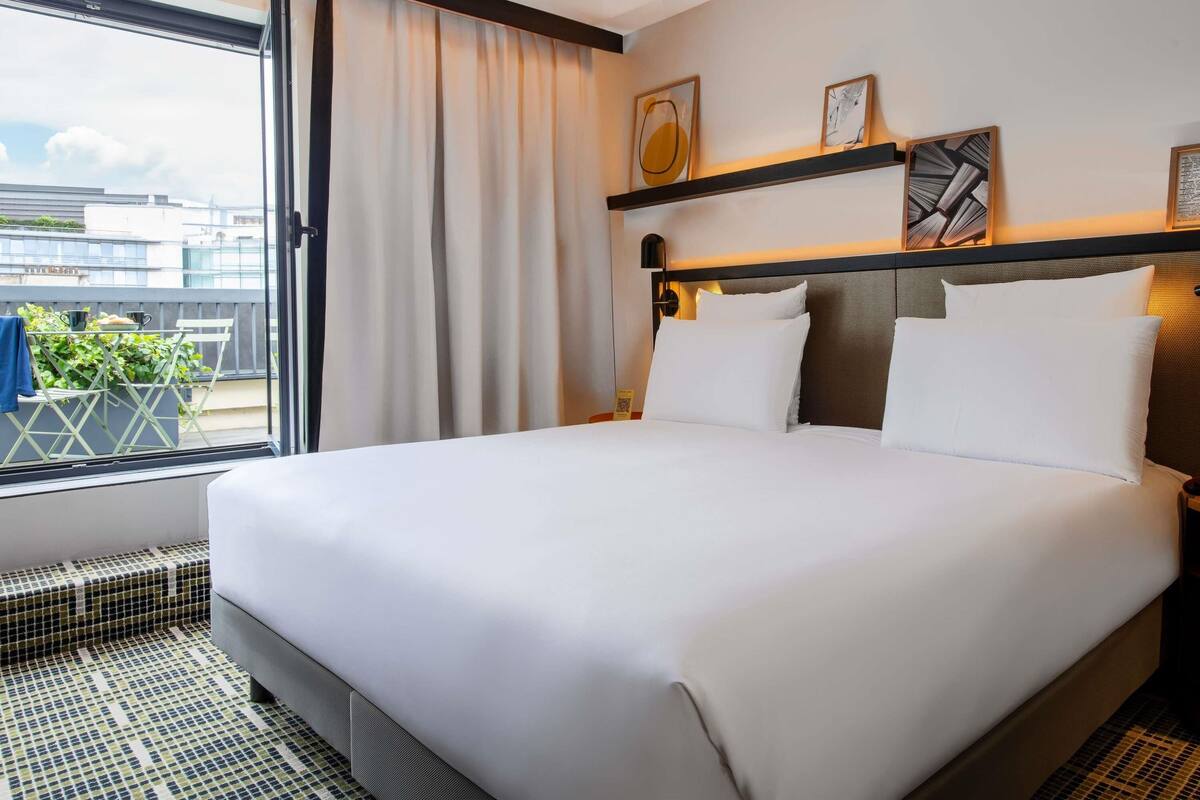 A hotel room with a large bed, a window with curtains, a balcony with a table and chairs, and artwork on the wall.
