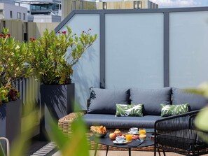 Un área de estar exterior moderna con sofá, cojines, una silla de mimbre y una mesa dispuesta con alimentos para el desayuno.
