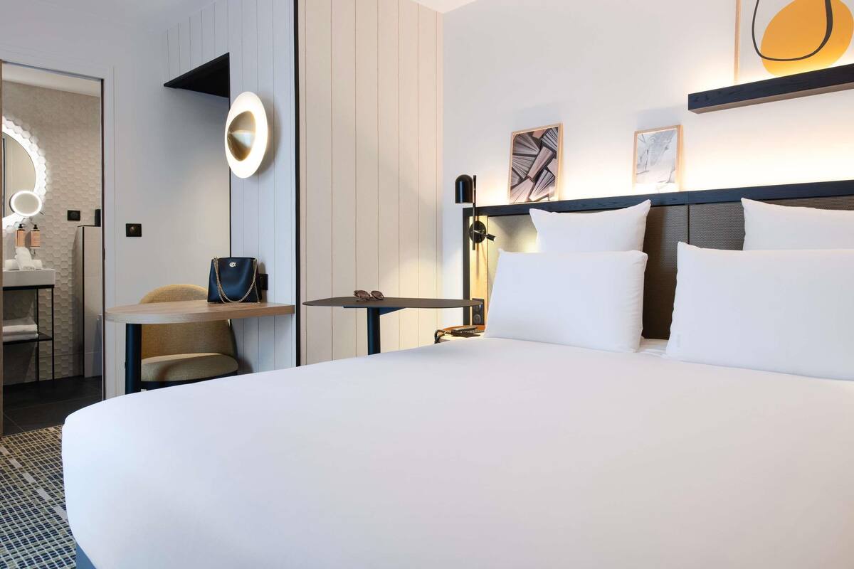 A modern hotel room with a large bed, a desk with a chair, a vanity with a mirror, and a wall-mounted lamp.