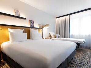 Ein Hotelzimmer mit einem großen Bett, einem Nachttisch, einer Lampe, einem Sessel und einem Fenster mit Vorhängen.