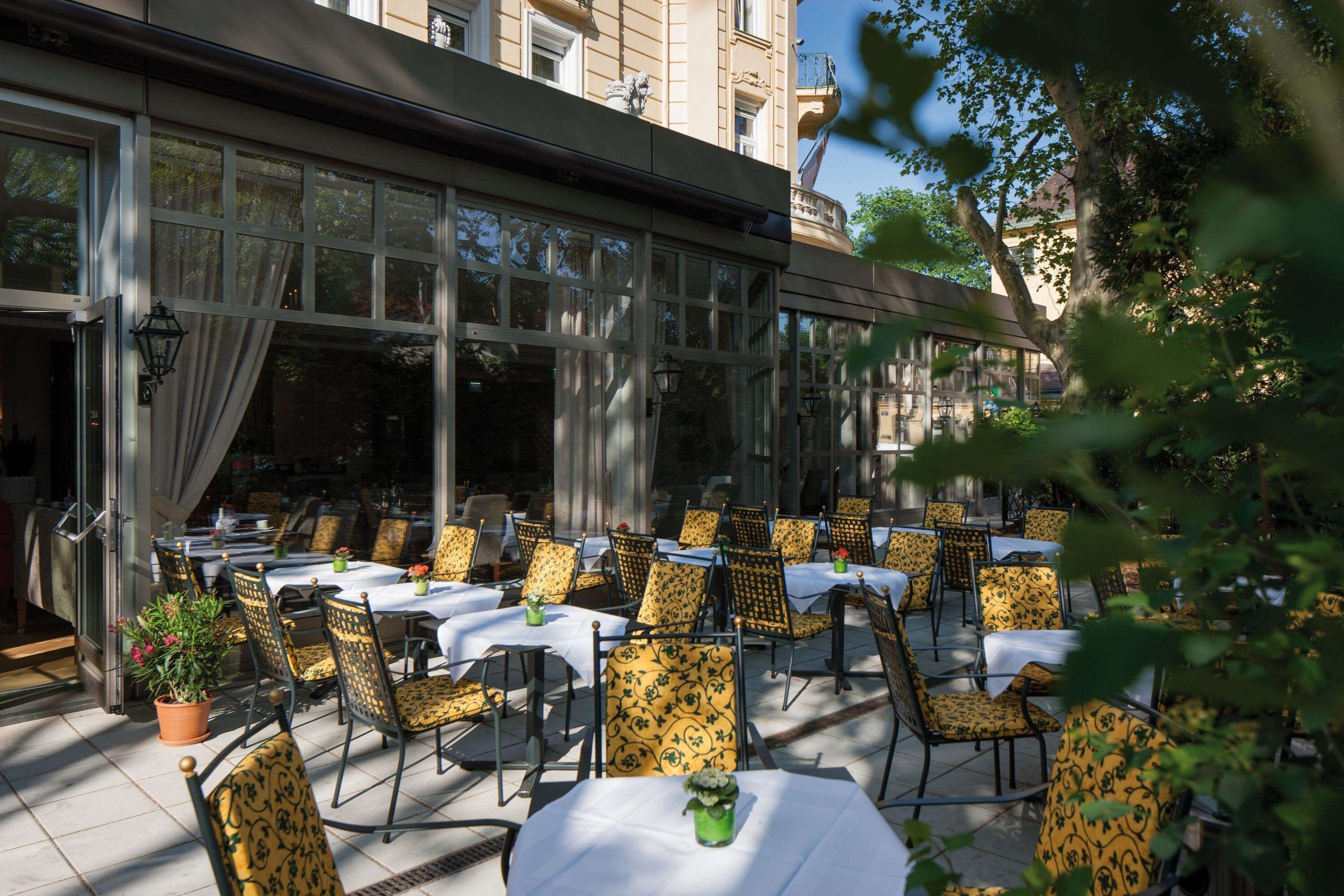 An outdoor seating area with tables and chairs, large windows, and potted plants.