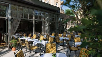An outdoor seating area with tables and chairs, large windows, and potted plants.