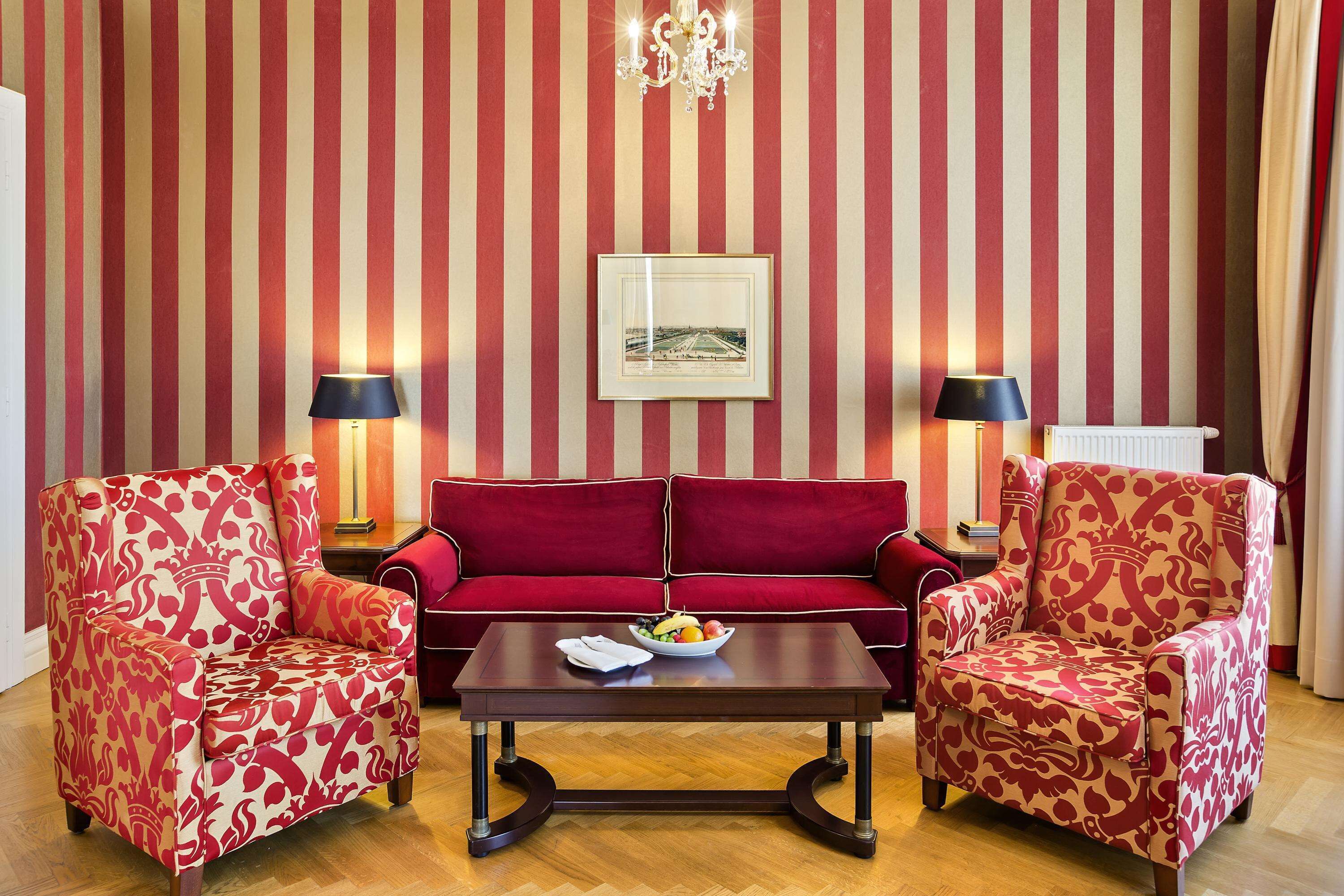 A room with striped wallpaper, a red sofa, two patterned armchairs, a coffee table with fruit, and two floor lamps.