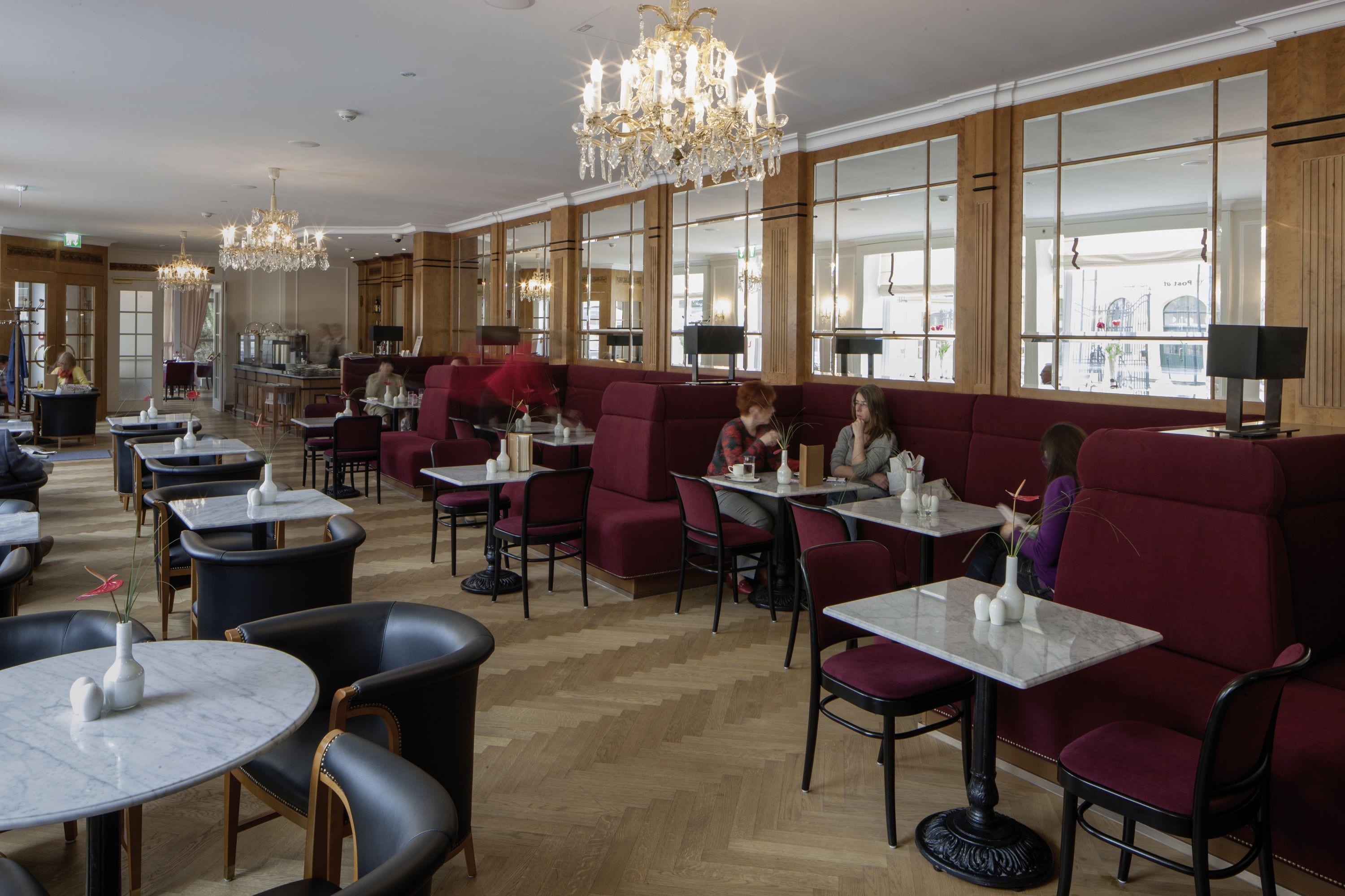A spacious restaurant with high ceilings, chandeliers, and large windows offering views of the street.