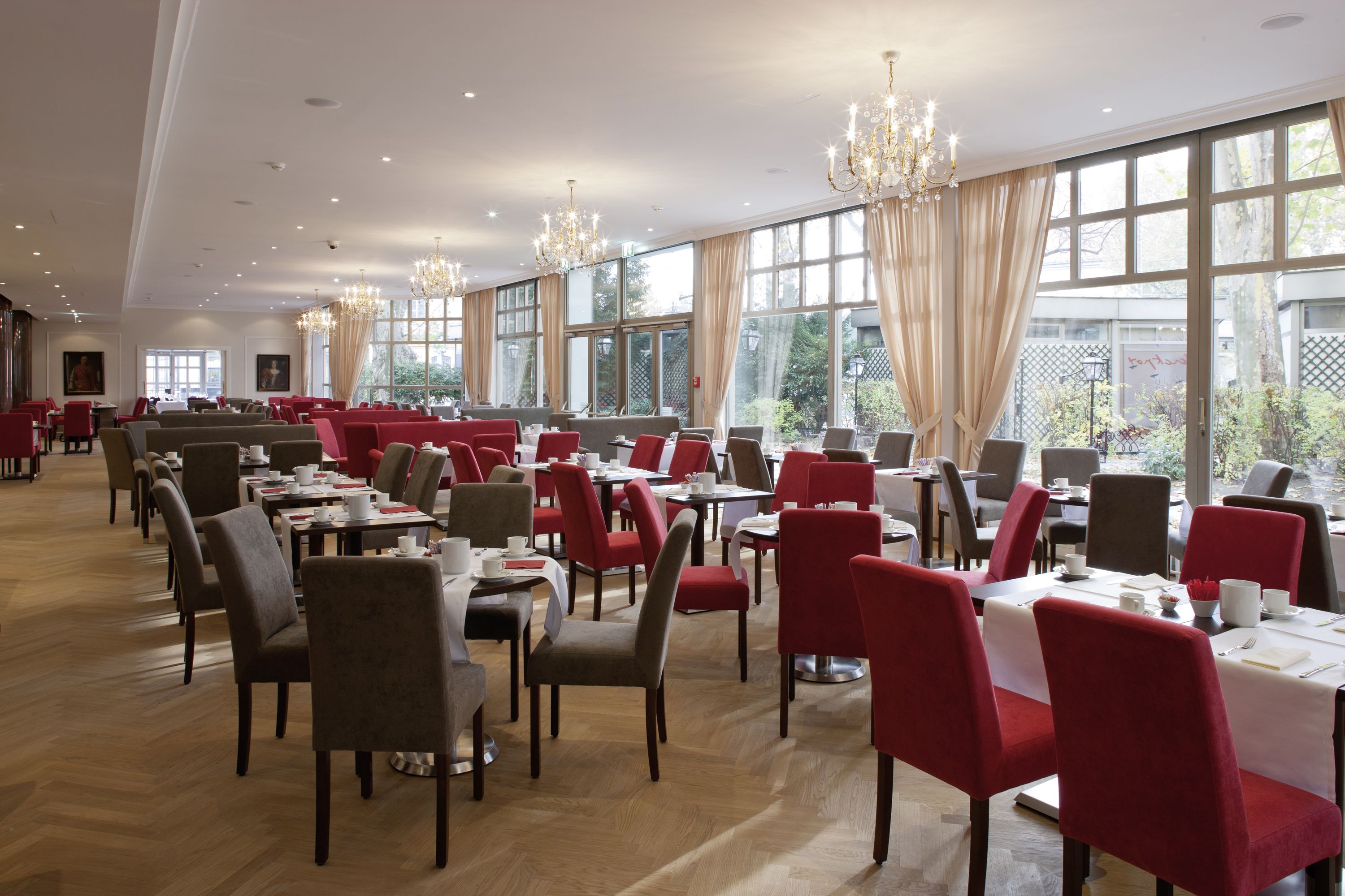 A spacious dining area with multiple tables and chairs, large windows with curtains, and chandeliers.