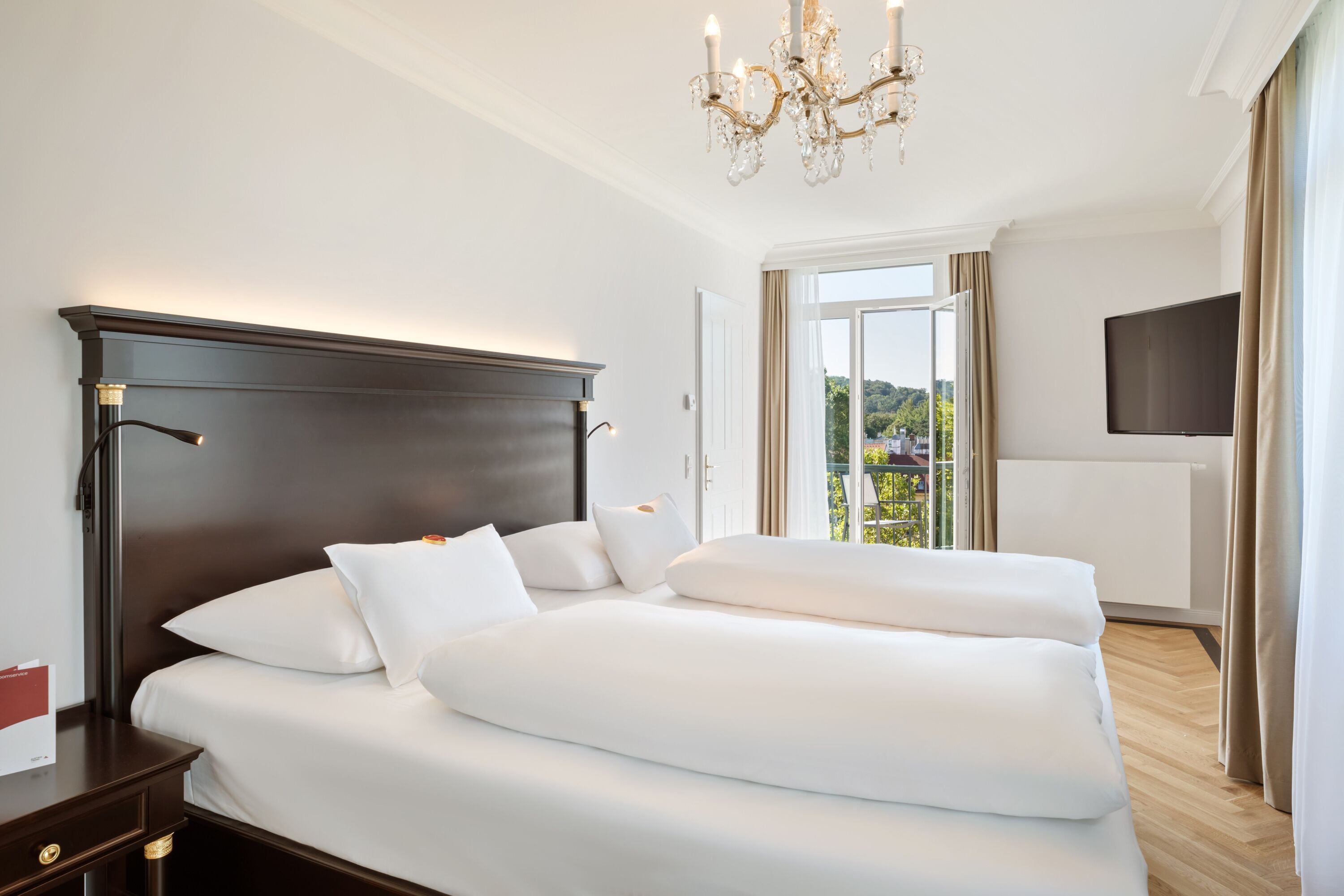 A hotel room with a large bed, a dark wooden headboard, a chandelier, a flat-screen TV, and a window with a view of greenery.
