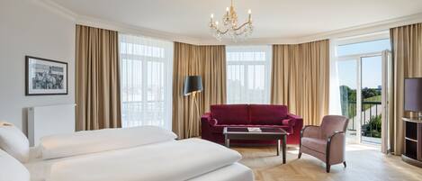 A hotel room with a large bed, a red sofa, a small table, and a TV. There are curtains, a chandelier, and a window with a view.