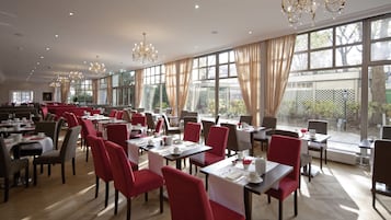 A spacious dining area with red chairs and tables, large windows, and chandeliers.