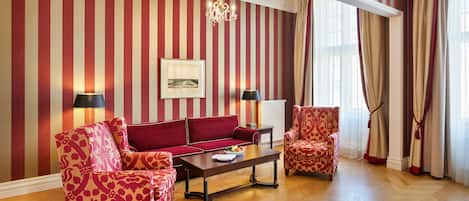A room with striped wallpaper, a chandelier, a red sofa, patterned armchairs, and a wooden floor.