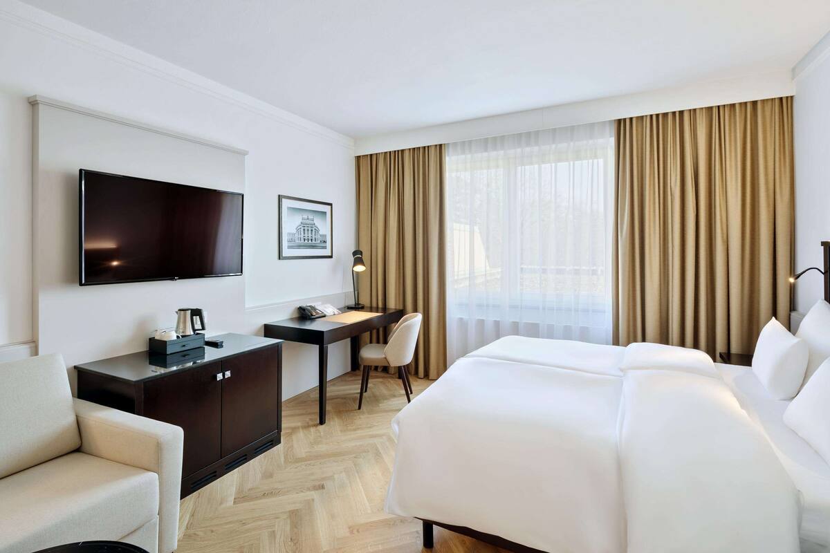 Ein Hotelzimmer mit einem großen Bett, einem Schreibtisch mit Stuhl, einem Fernseher und einem Fenster mit Vorhängen.