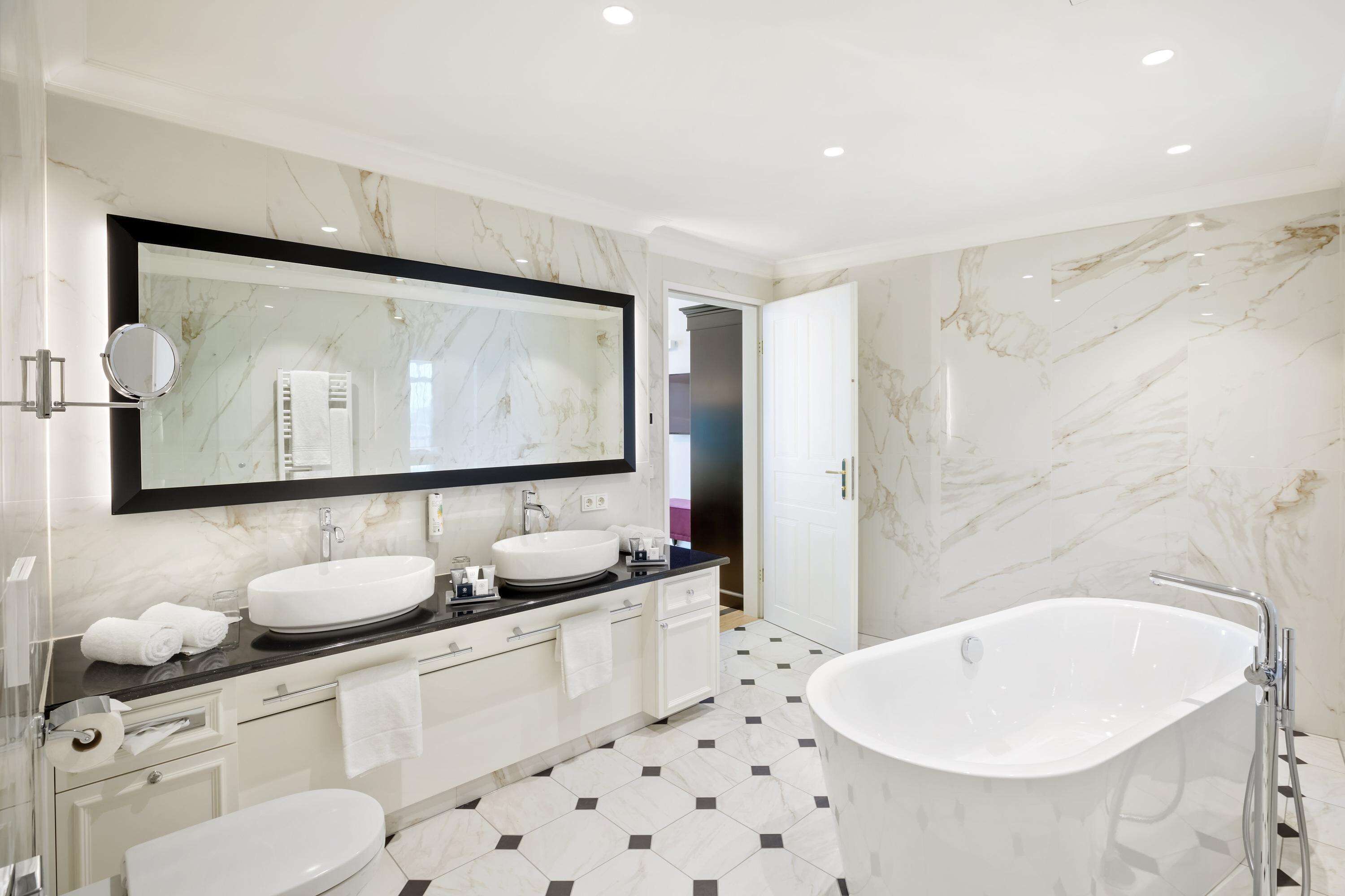 A modern bathroom with a large mirror, two sinks, a bathtub, and a toilet.