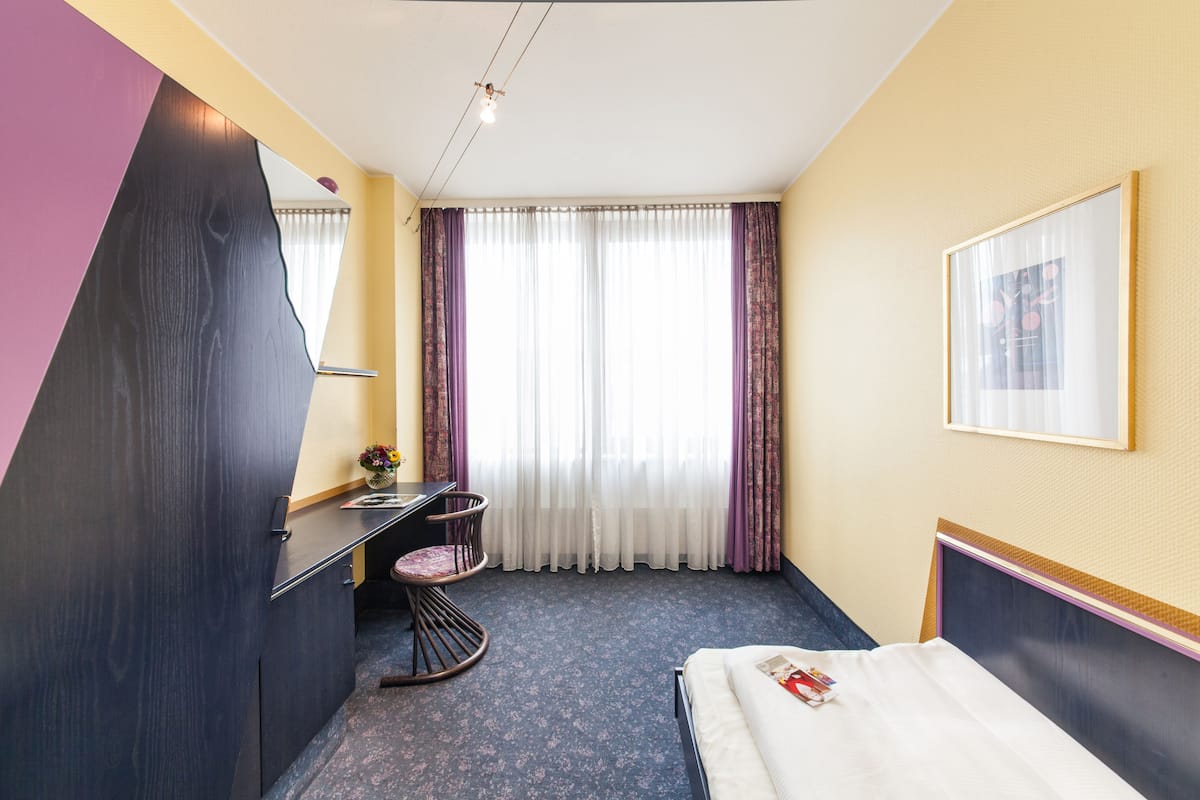 A hotel room with a bed, a desk, a chair, a large mirror, and a window with curtains.
