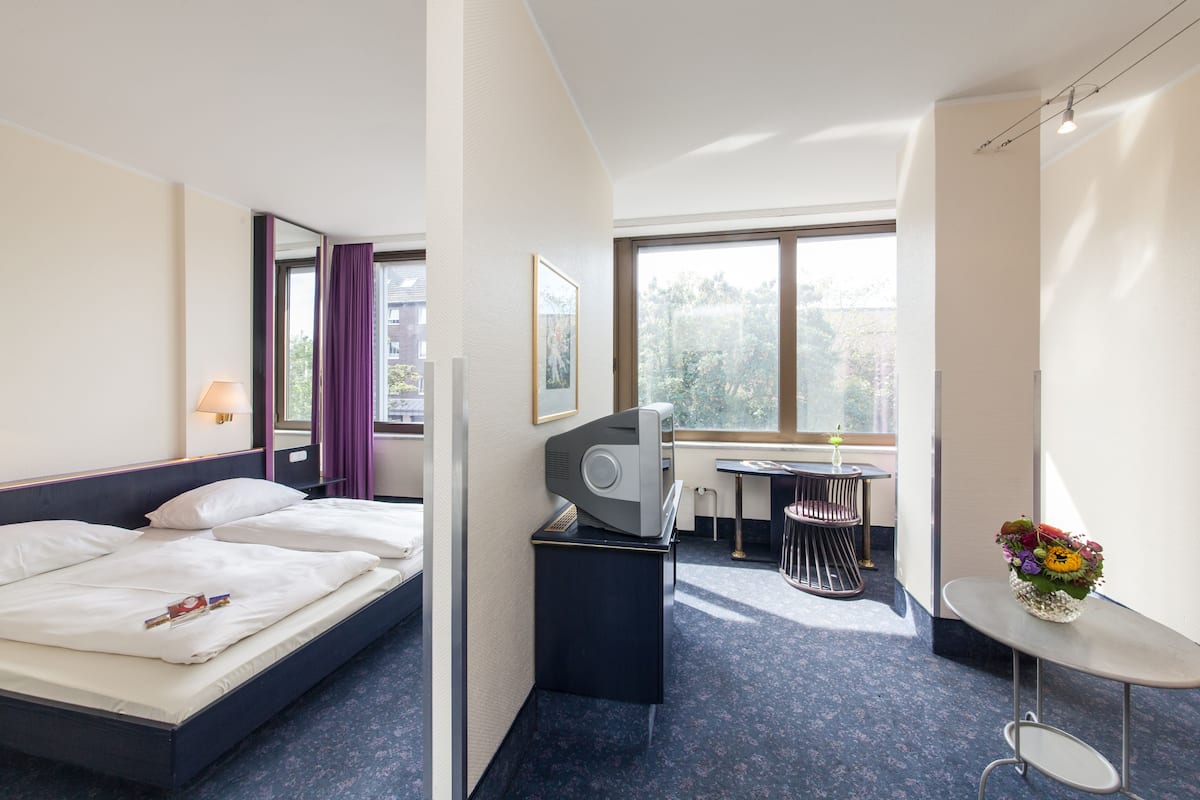 A hotel room with a bed, a desk, a chair, a small table with a vase of flowers, and a window with a view of trees.