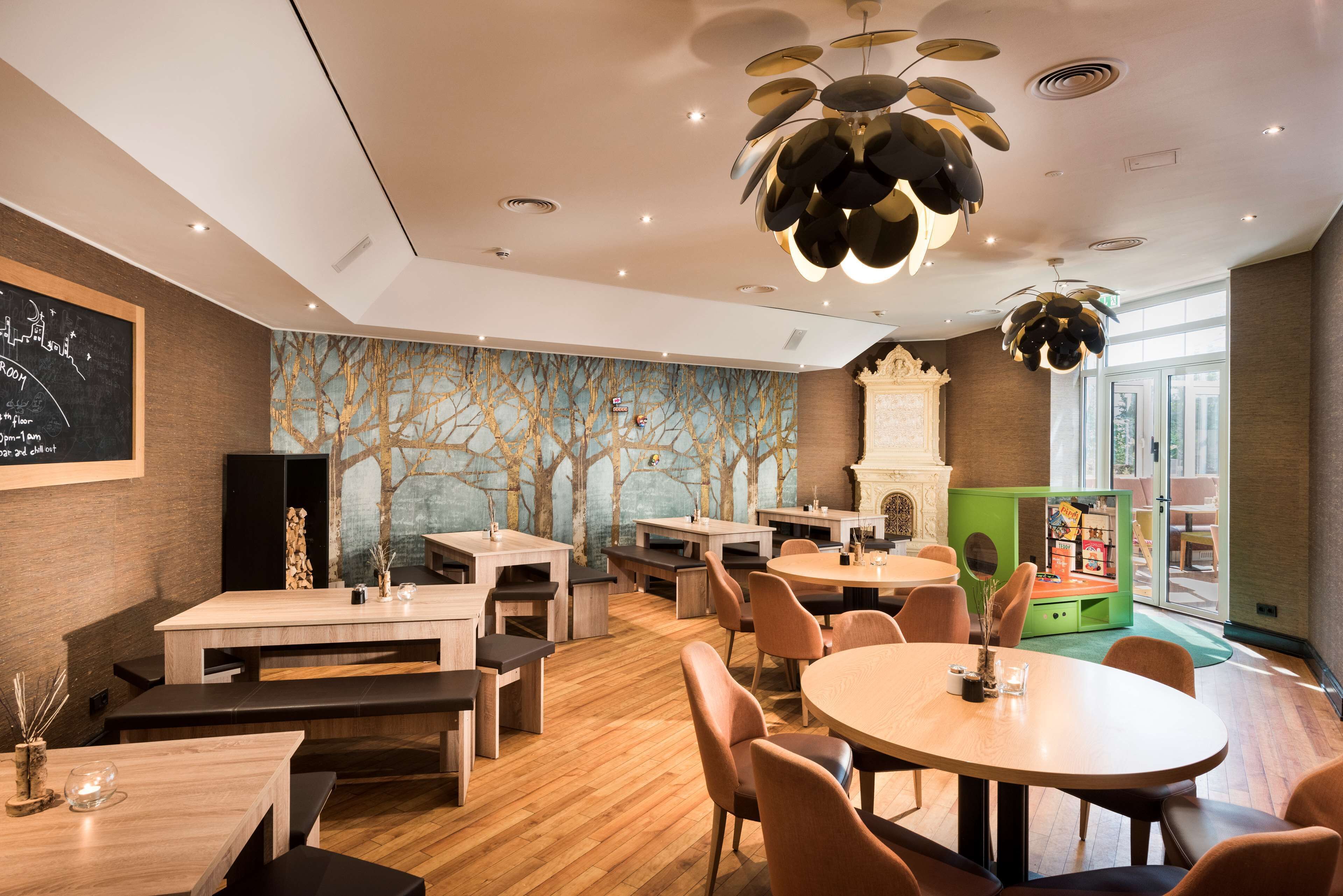 Un restaurante moderno con mesas y bancos de madera, un mural de árboles en la pared y un área de juegos verde para niños.