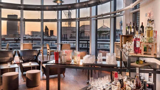 A modern bar area with a panoramic city view, leather seating, and a variety of bottles and glasses. - Scandic Berlin Kurfürstendamm (Berlin)