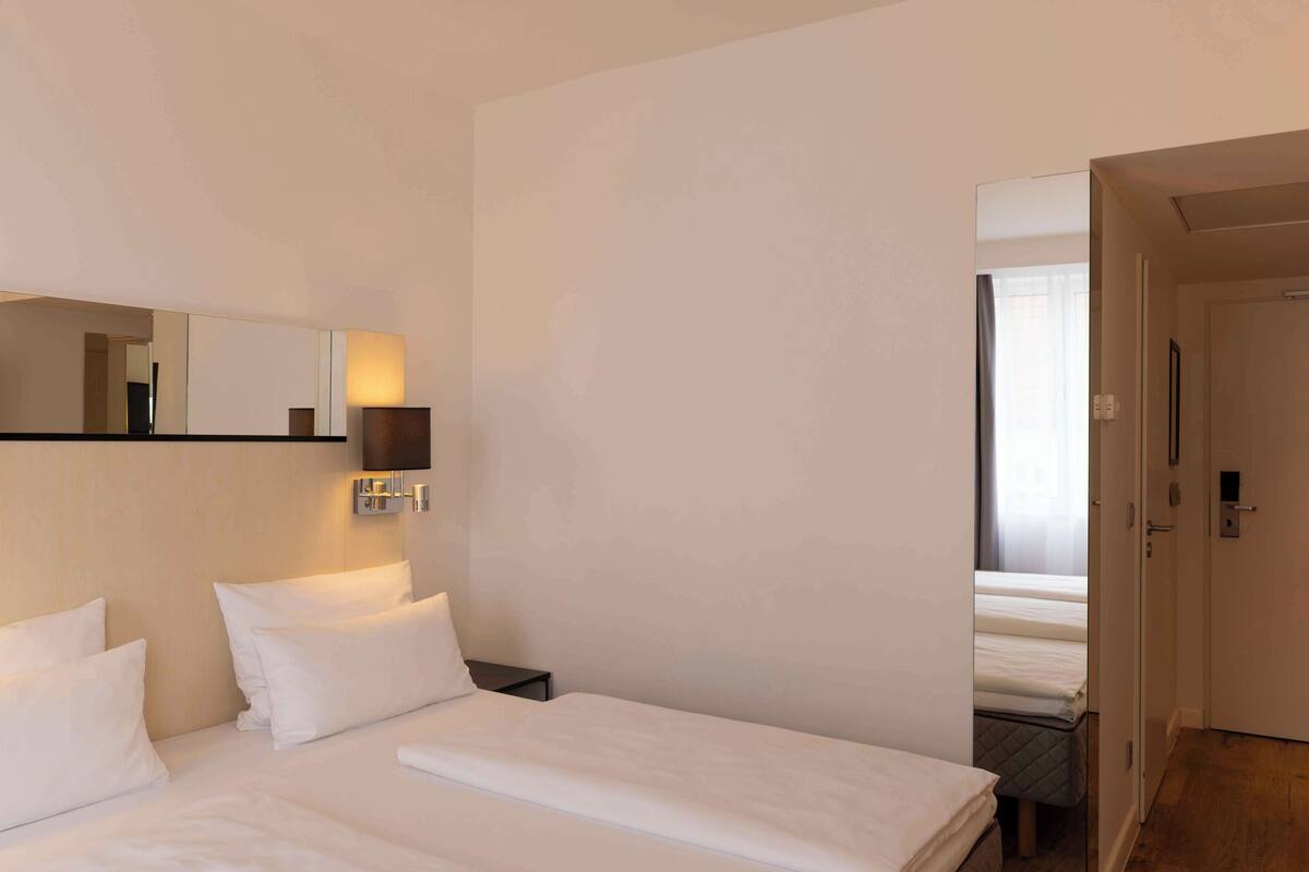 A hotel room with a large bed, a bedside table, a wall-mounted lamp, and a door leading to another room.