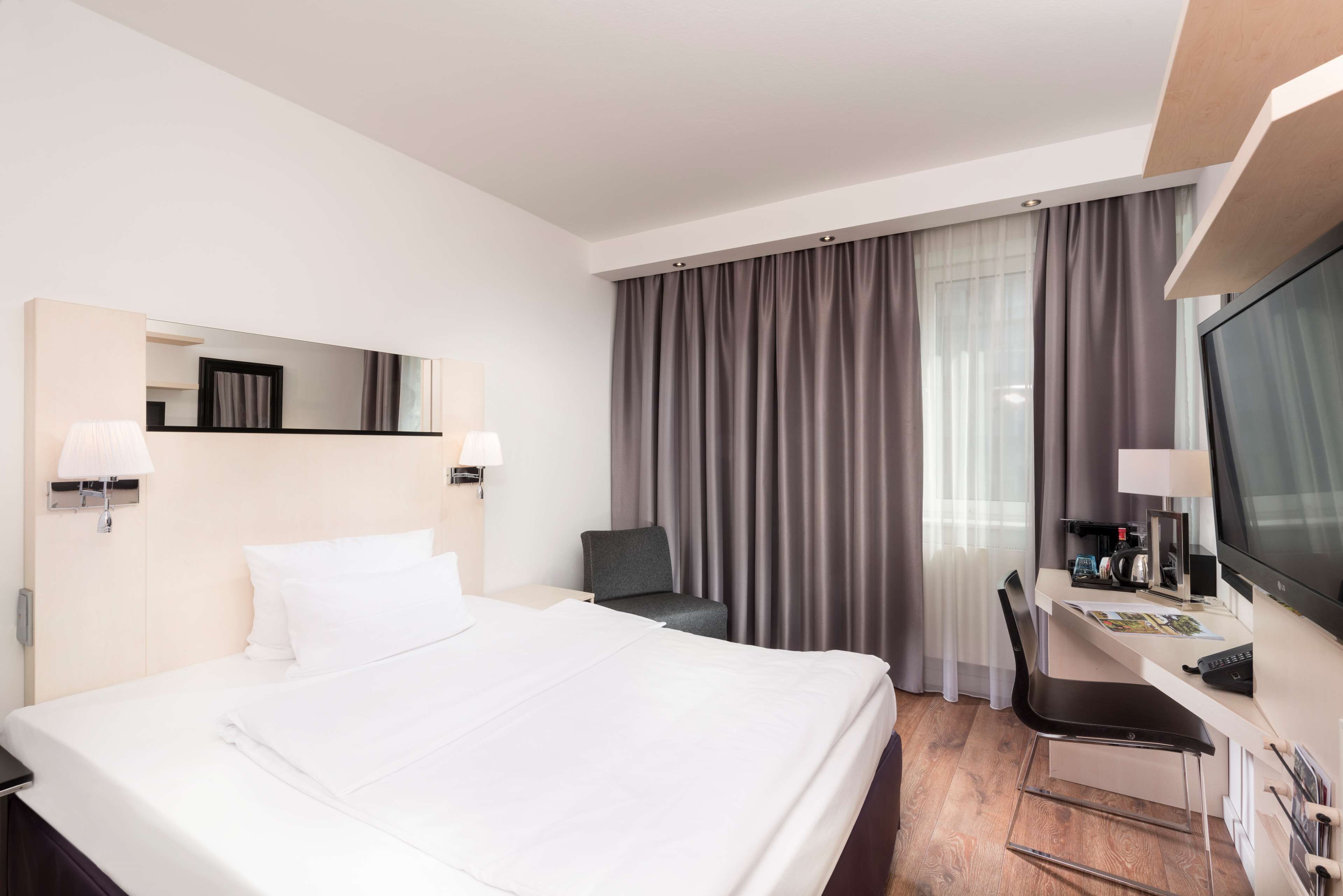 Habitación de hotel con una cama grande, un escritorio con silla, un televisor y una ventana con cortinas.